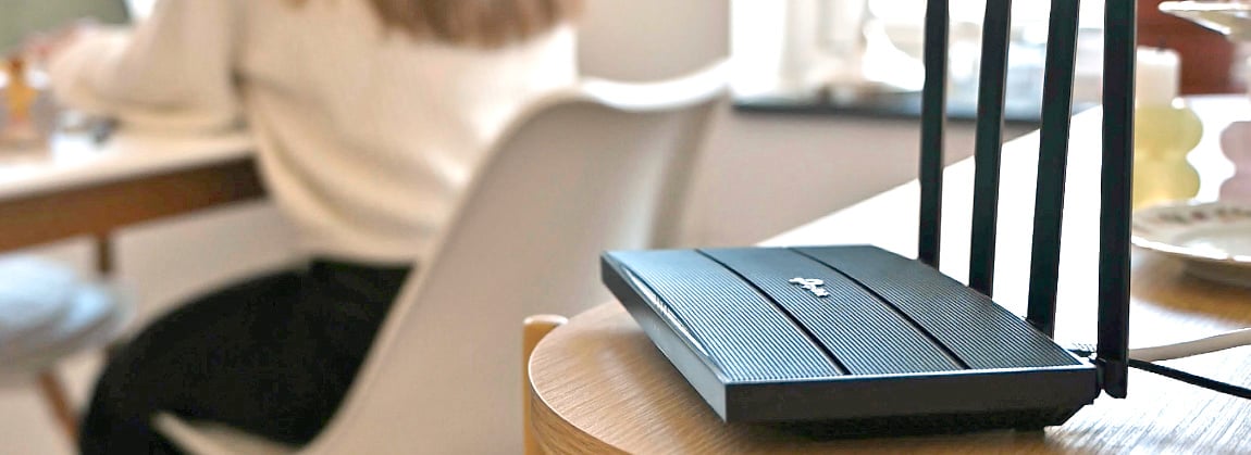 Svart WiFi-router med fyra antenner står på runt träbord i hemmiljö med kvinna vid dator i bakgrunden.