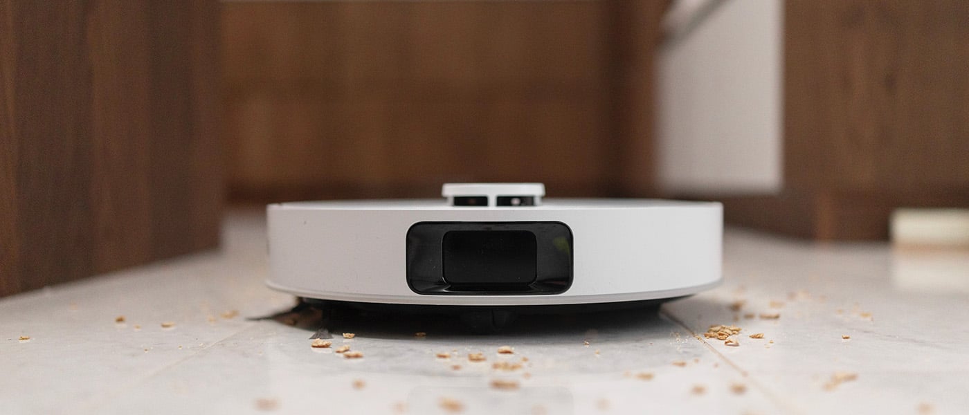 Närbild framifrån på en vit robotdammsugare som dammsuger upp smulor från ett kakelgolv.