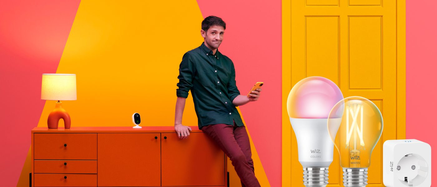 Man håller i en smartphone omgiven av WiZ smarta produkter – glödlampor, kamera och eluttag – mot orange och rosa bakgrund.