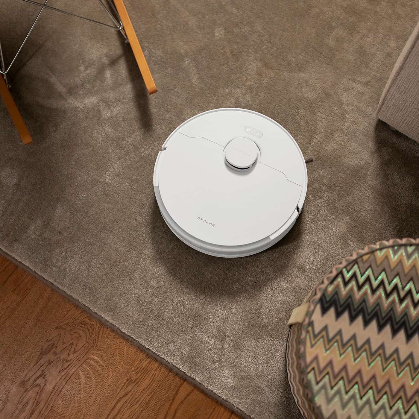 Vit robotdammsugare från Dreame kör på en brun matta mellan möbler i ett vardagsrum.