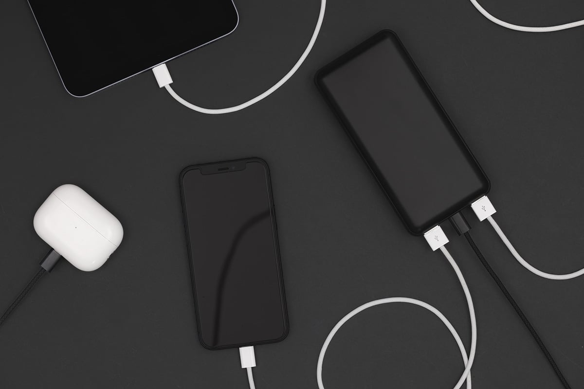 Svart mobil, surfplatta, powerbank och vita hörlurar, alla anslutna med laddkablar på svart bakgrund.
