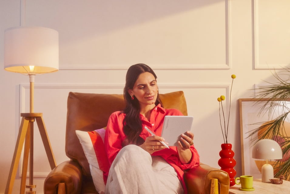 Kvinna i röd skjorta sitter i fåtölj och skriver på en surfplatta med penna i ett varmt inrett rum.