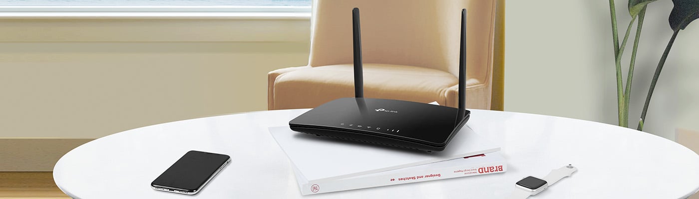 Svart router med två antenner placerad på en bok, bredvid en smartklocka och en smartphone på ett vitt bord.