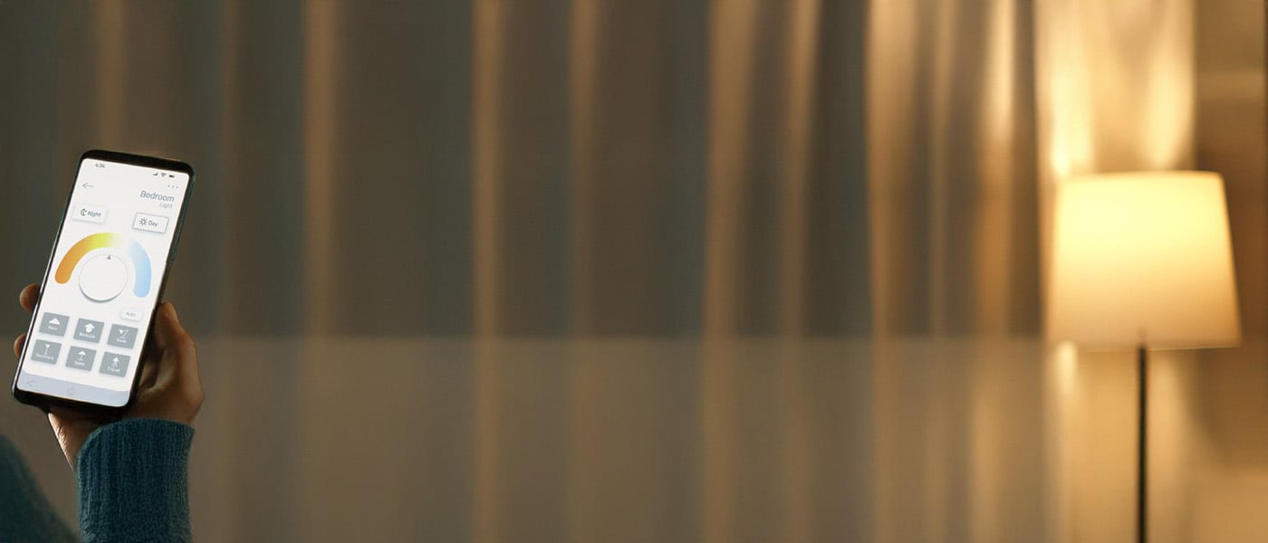 Hand som styr varmvit belysning med en mobilapp i ett dämpat rum med golvlampa i bakgrunden.