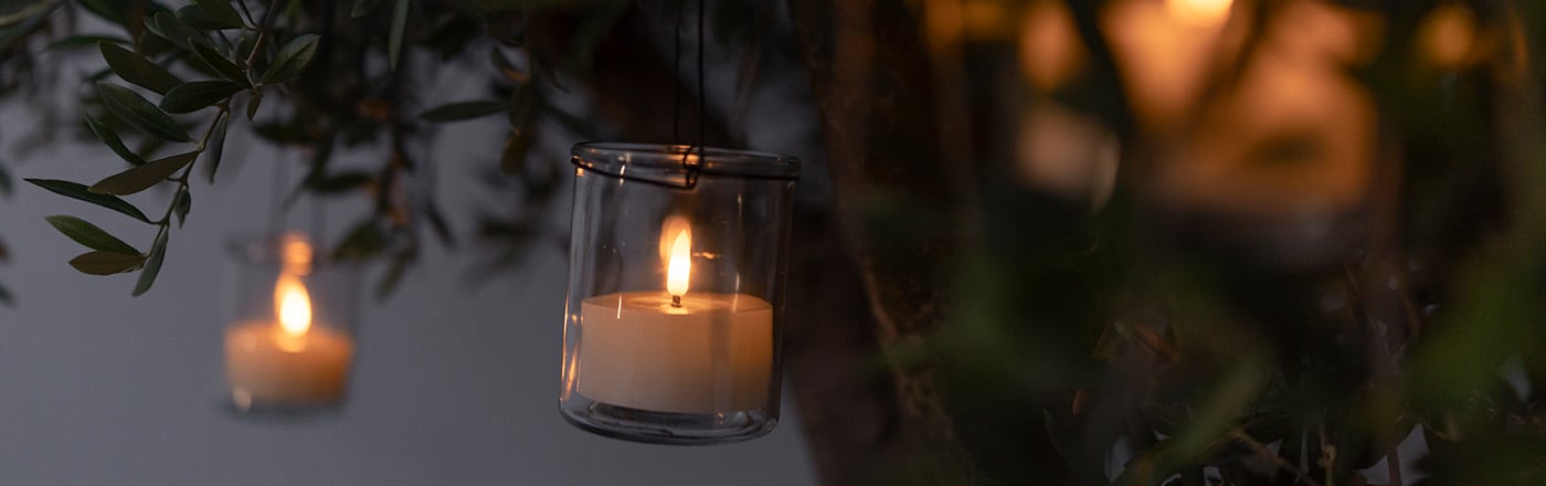 Brinnande värmeljus i små glaslyktor hängande i trädgren bland gröna blad i skymning.