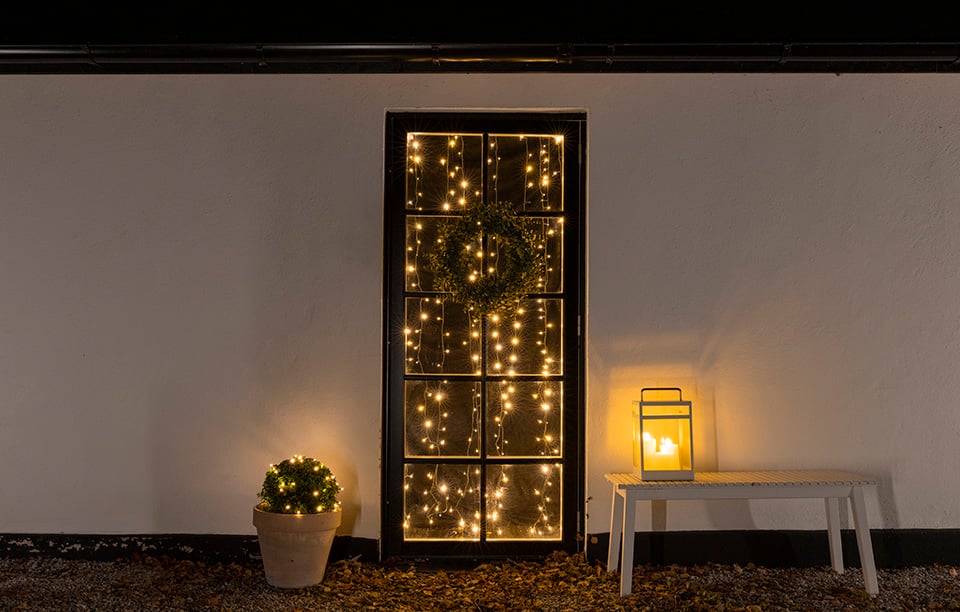 Upplyst trädstam med ljusslinga, tre lyktor med LED-ljus och filtar på en altan i kvällsbelysning.