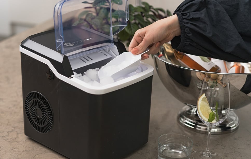 Svart ismaskin med öppen lucka, någon skopar upp isbitar bredvid dryckeskyl och glas med citron.