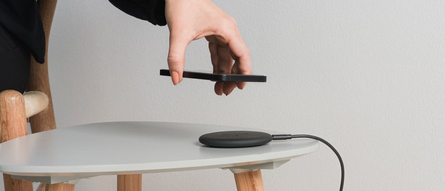 Hand placerar en svart smartphone ovanför en rund, svart trådlös laddare på ett vitt bord.