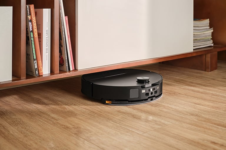 Svart robotdammsugare kör in under en låg bokhylla på ett ljust trägolv i hemmiljö.