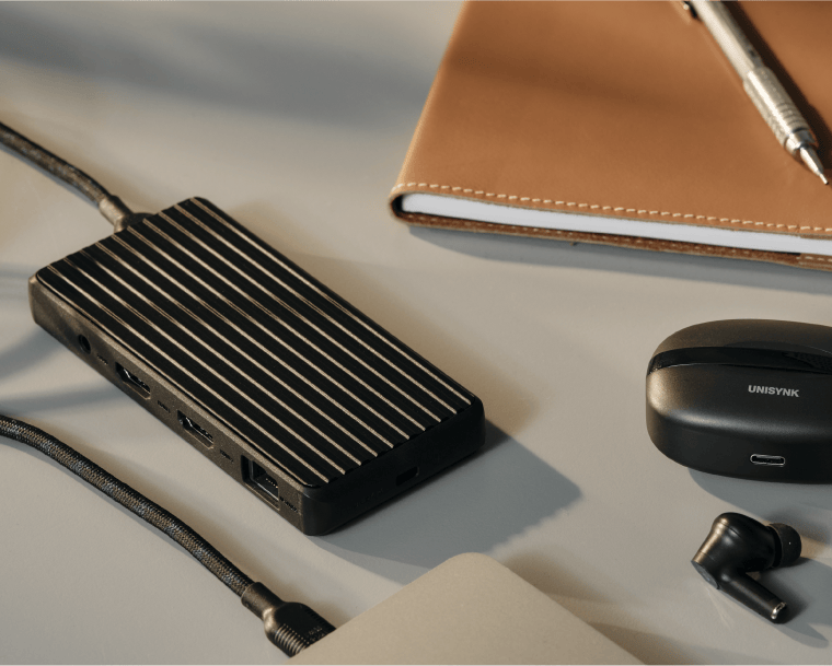 Svart Unisynk-hubb med ribbad yta på skrivbord, ansluten till flera kablar, omgiven av anteckningsbok och hörlurar.