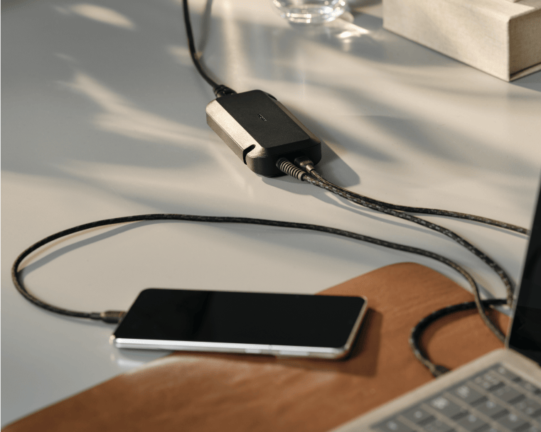 Unisynk-laddare på skrivbord, ansluten mellan bärbar dator och mobiltelefon med flätade kablar.
