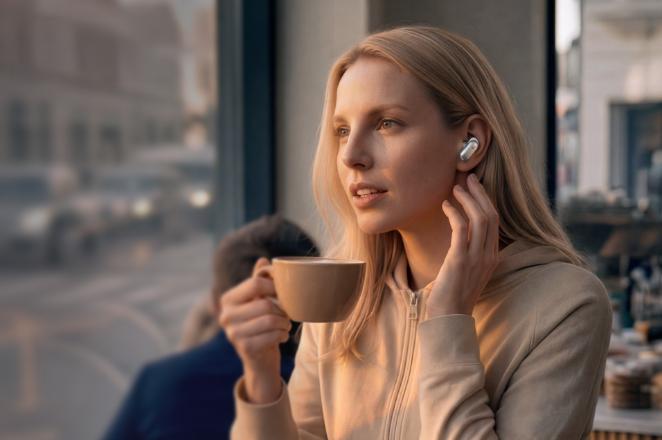 Kvinna med trådlösa Soundcore-hörlurar håller en kaffekopp vid ett caféfönster.