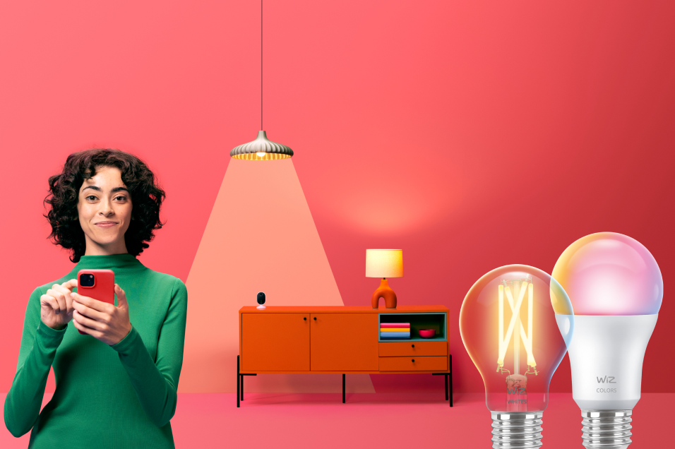 Smart belysning från WiZ, inklusive LED-lampor, ljusslinga och kontakt, med avslappnad man på grön soffa i orange miljö.