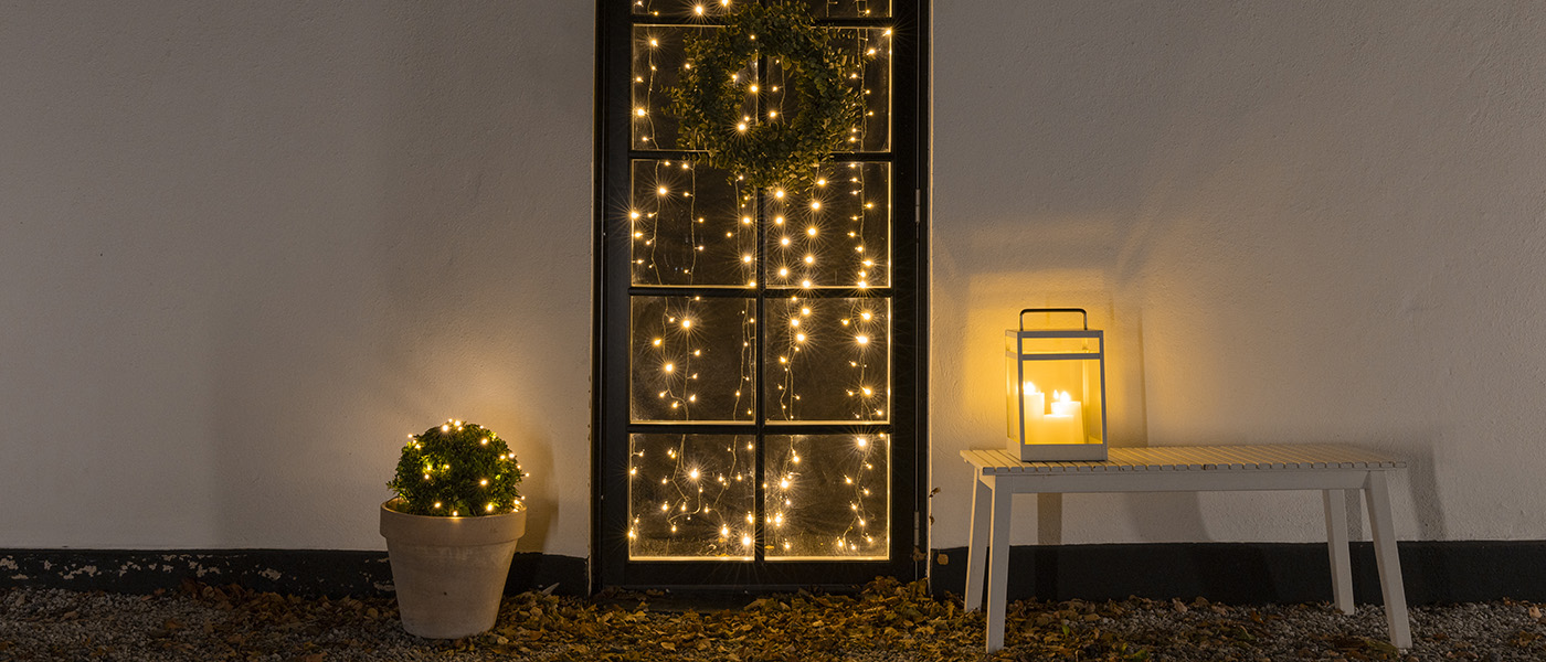 Svart dörr med ljusslingor och grön krans, kruka med ljusslingor till vänster och lykta med LED-ljus på vit bänk till höger.