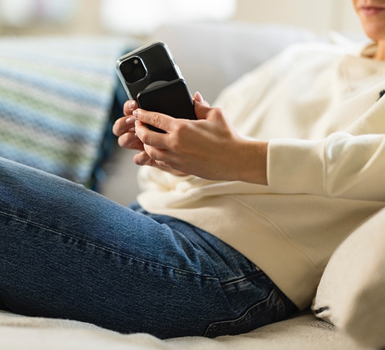 Smarttelefon med MagSafe-deksel og MagSafe-powerbank festet på baksiden, holdes i hånden av person som sitter i sofa i hjemmemiljø.