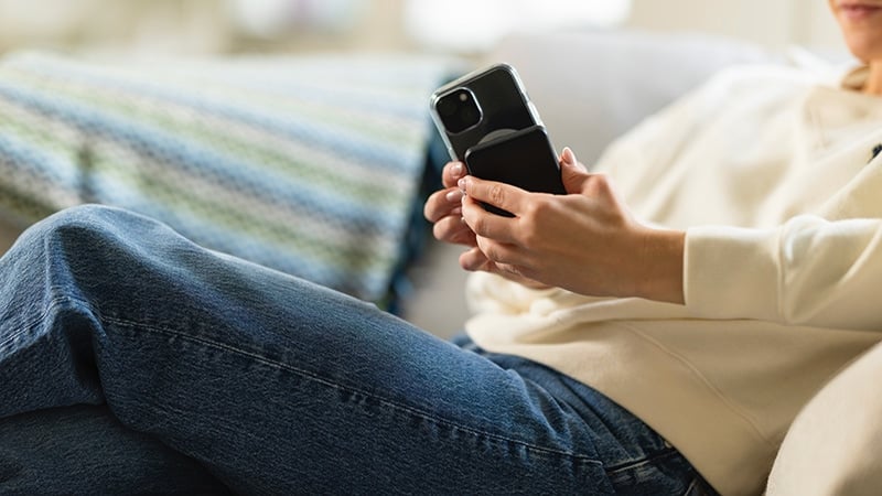 Smarttelefon med MagSafe-deksel og MagSafe-powerbank festet på baksiden, holdes i hånden av person som sitter i sofa i hjemmemiljø.