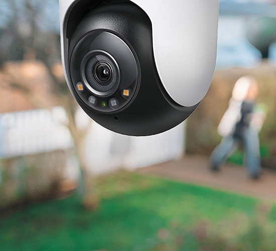 Närbild på en vit och svart övervakningskamera monterad utomhus med suddig trädgård och person i bakgrunden.