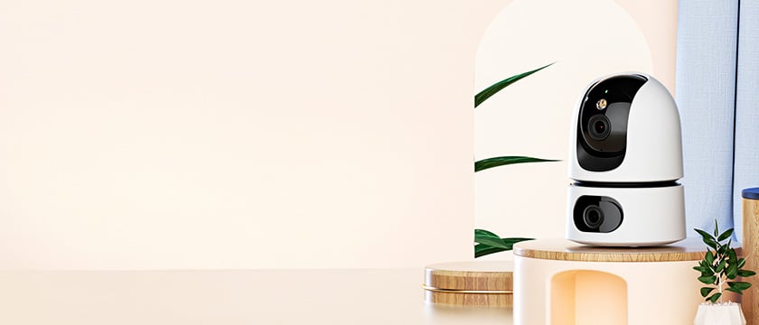 Hvit overvåkningskamera med svart front i avrundet design, stående på en treplattform i et stilrent interiør med grønne blader.