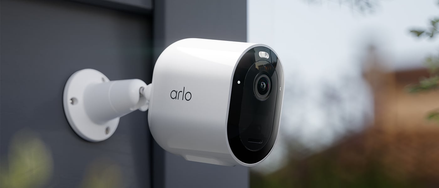 Hvit Arlo-overvåkningskamera montert på en mørk yttervegg, vendt utover for å overvåke området.
