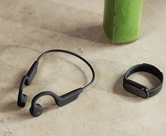 Svarta trådlösa benledningshörlurar och ett svart aktivitetsarmband ligger på ett ljust bord intill en grön smoothieflaska.