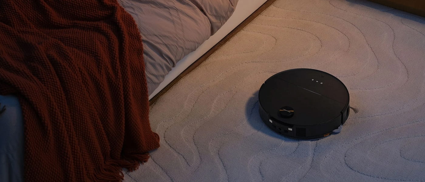 Svart robotdammsugare på ljus matta i sovrumsmiljö, intill säng med rostfärgad filt och lila täcke.