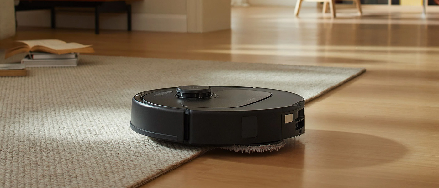 Svart robotdammsugare i drift, kör från ljus trämatta upp på en beige matta i ett ljust vardagsrum.