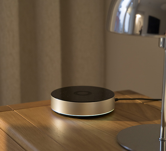 Rund smart hem-hub med svart topp och metallram, placerad på ett träbord med svagt lysande LED-ring i botten.